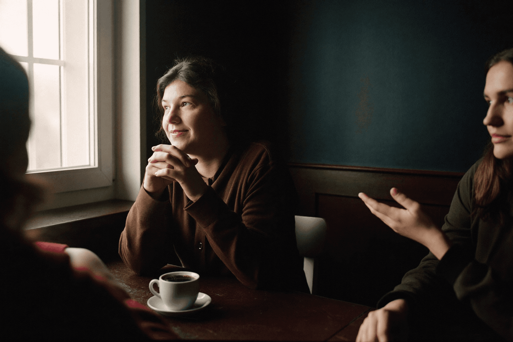 A person sitting at a table by a window with a mug in front of them, someone gesturing beside them.