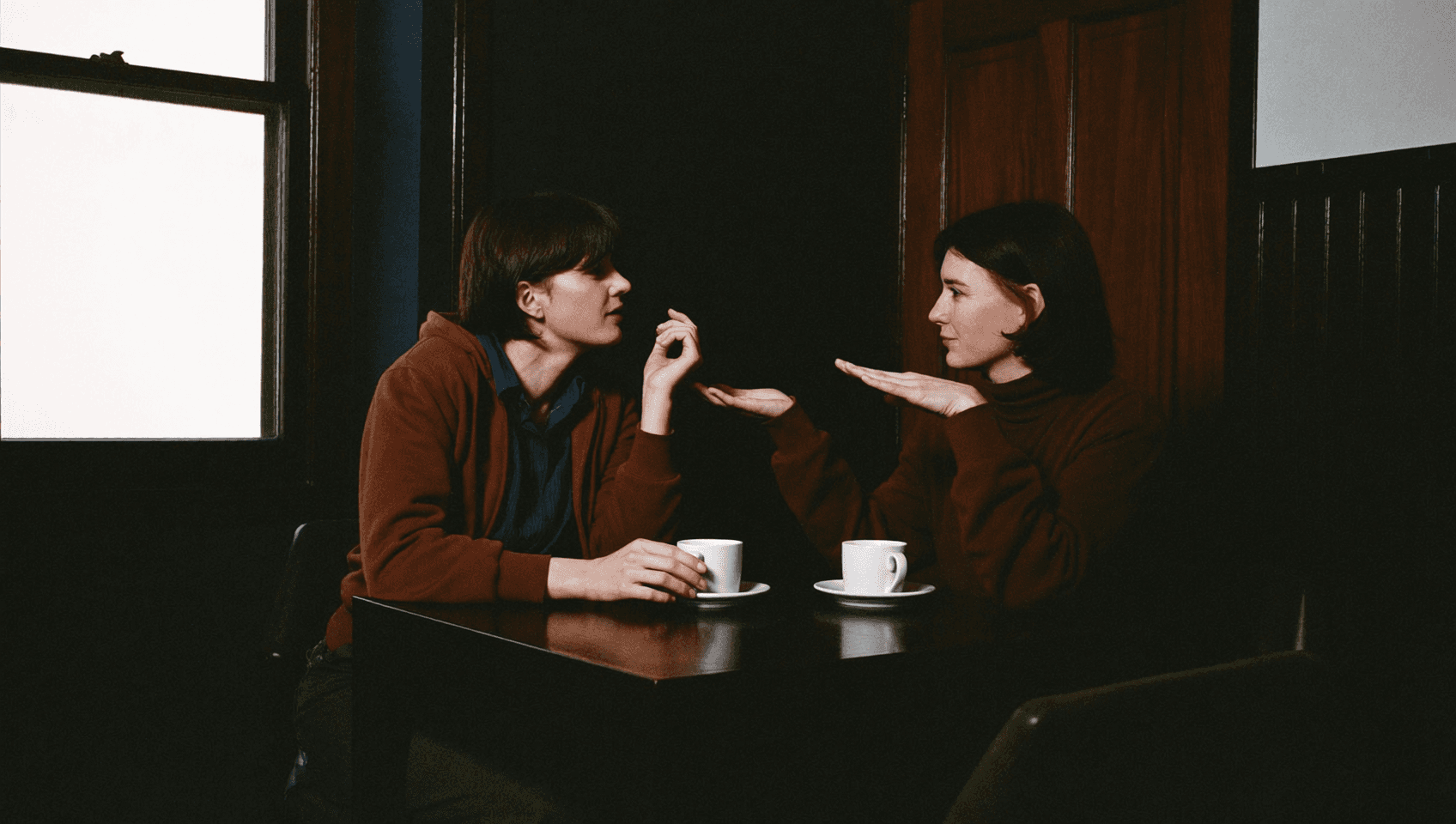 Two people at a table, one speaking with a raised hand while the other listens.
