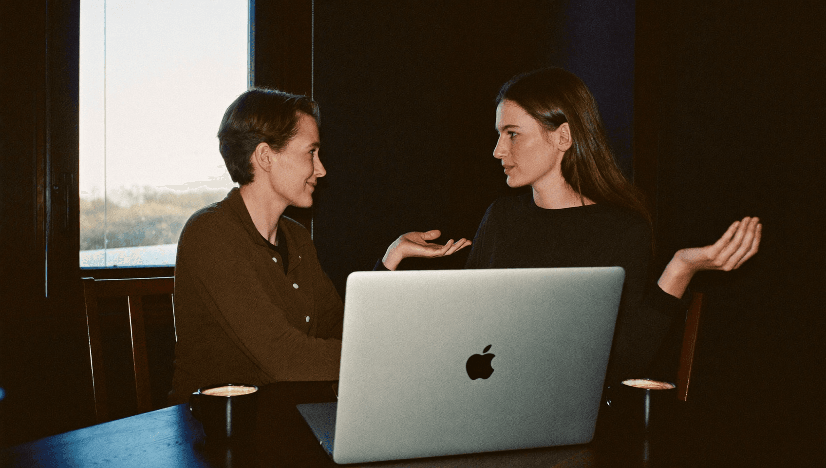 Two people talking over an open laptop, seen from behind the lid.
