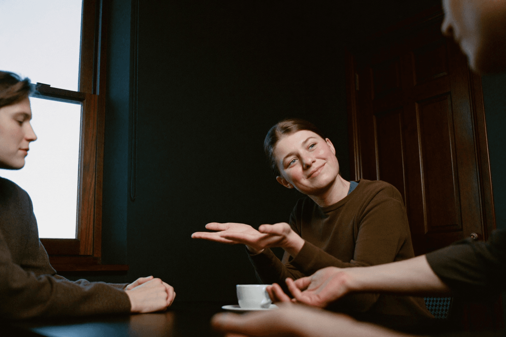 Several people at a table, the one in the middle of the frame looking at the others, a cup in front of them.