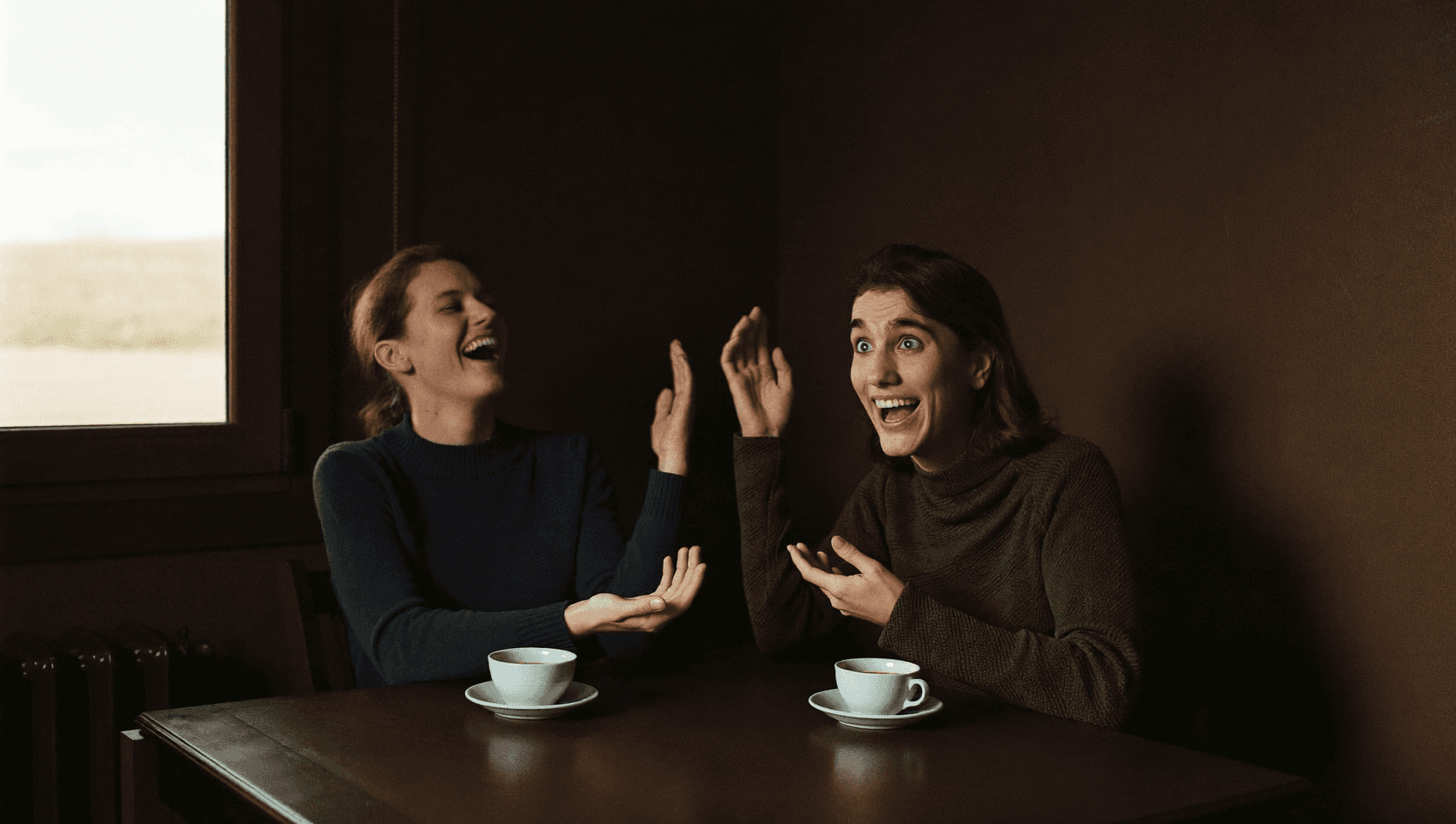 Two people at a table laughing out loud, both with their hands in the air.