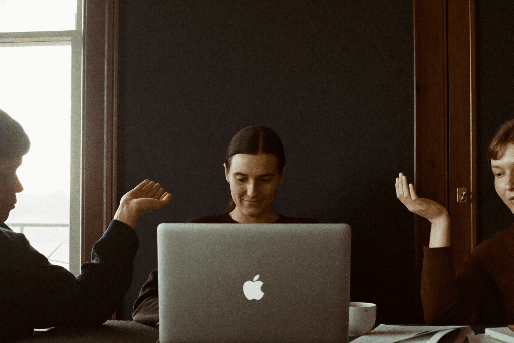 An open laptop in the foreground, someone looking at the screen behind it and a raised hand beside them.