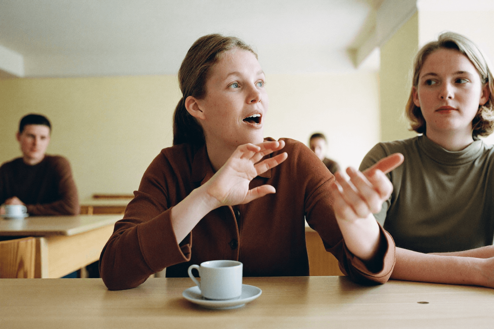 A person mid-sentence with a raised hand, someone listening beside them.