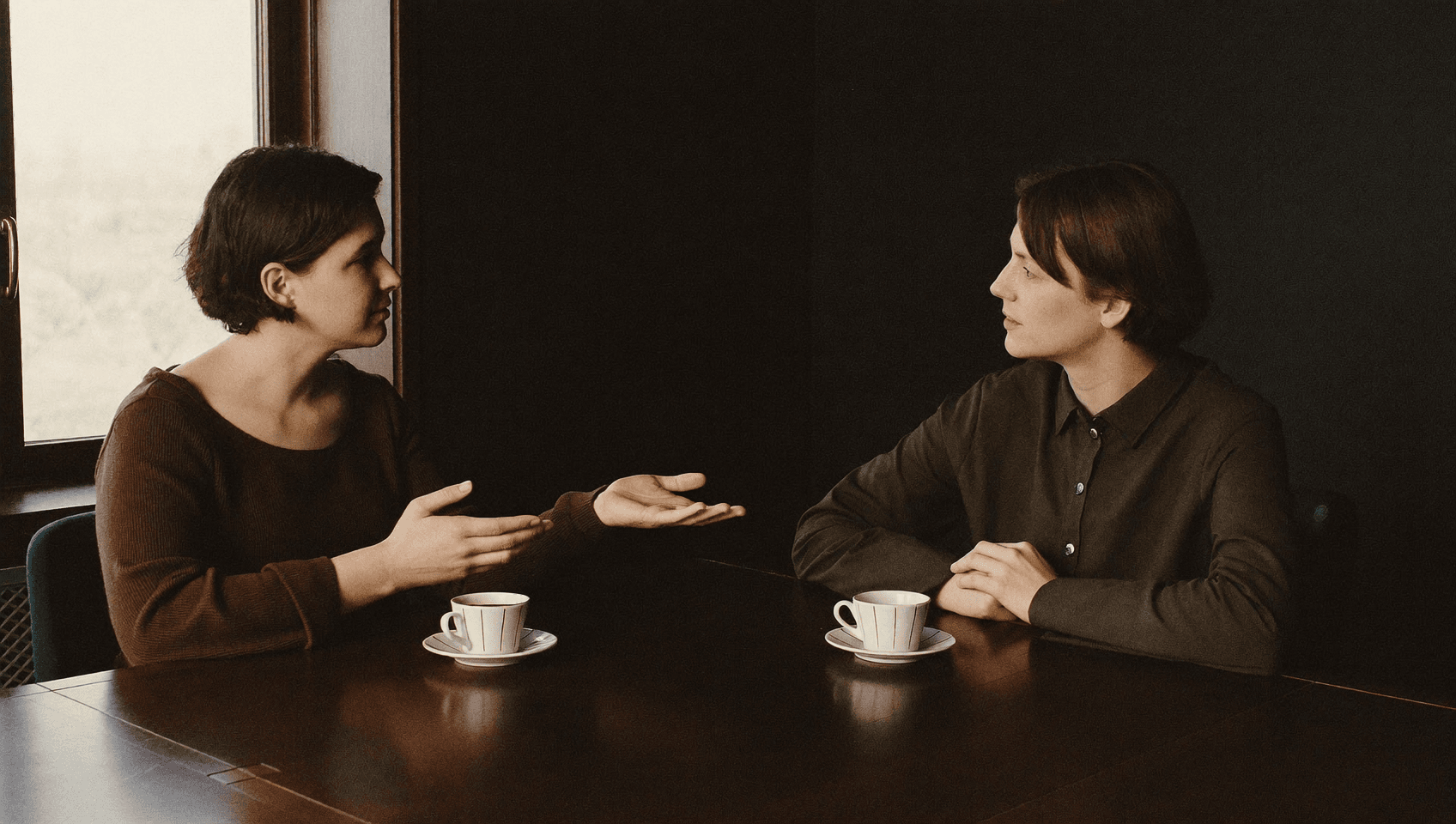 Two people facing each other across a table, one explaining with open hands, cups beside them.
